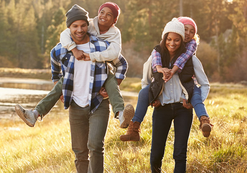 Young family of four out hiking in the woods. The two adults are giving the children piggy back rides. The family is dressed warmly and they are all smiling.
