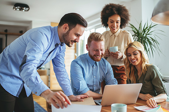 Four adults in business casual dress gather around a worktable and are looking at a laptop computer. There are two men on the left and two women on the right. They are all smiling and looking at the screen.