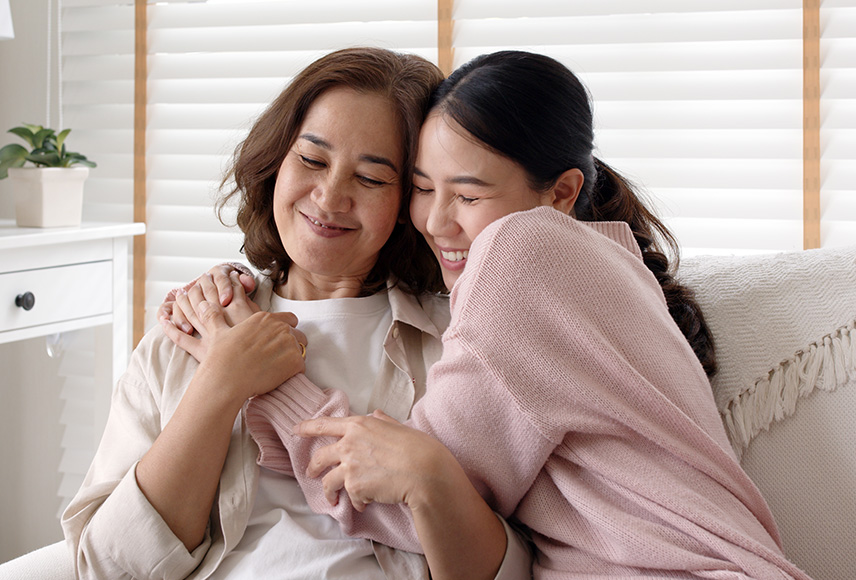 A woman in her 50s is seated on a couch next to her teenage daughter. The daughter has her arms around her in a side hug. The mother is reaching her arms up to grasp the front arm of the daughter. Both of their faces are scrunched up in a smile and not making eye contact with the camera.