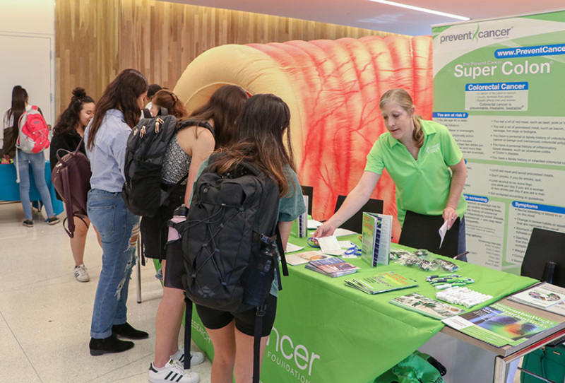 A Fondation staff person is behind a behind a table at a health fair wearing a bright green shirt. She is handing out materials to a group of people.