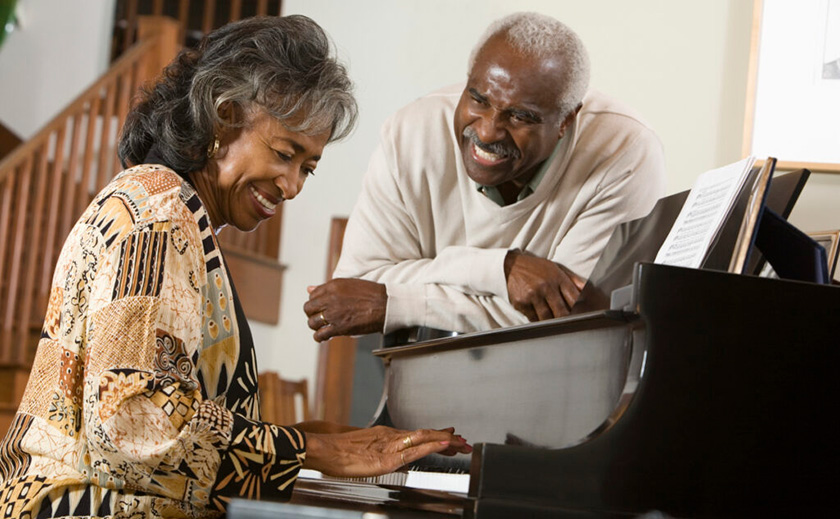 A close portrait of a senior couple at a piano. A woman is seated in front of the piano and is playing. The man is to her left and leaning in and on the piano. Both are smiling.