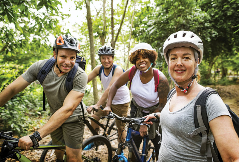 Four diverse adults in their 40s and 50s are out in the woods mountain biking. They are facing the camera with smiles and wearing helmets.