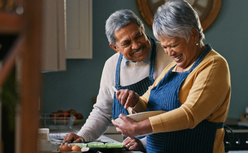 A senior man and woman are standing side by side in a kitchen preparing a meal. Both are wearing aprons and grinning broadly.