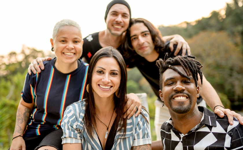 An intimate portrait of a diverse group of people in their 20s and 30s. In the front row is a white woman with long brown hair and a Black man with short dreadlocks and a goatee. The back row is a white woman with a blond buzz cut, a Hispanic man wearing a beanie and white man with shoulder-length brown hair. Everyone is smiling and facing the camera.
