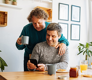 A white man in his 50s is standing, holds a cup of coffee and rests his other hand on the should of his husband. The other man is seated at a table hold a phone. Both are looking down at a the phone screen.