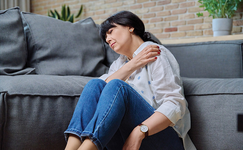 Image of a white woman in her 50s with dark hair sitting on the floor in front of a gray couch. She is reaching across her body to touch her left should and has a grimace of pain on her face.