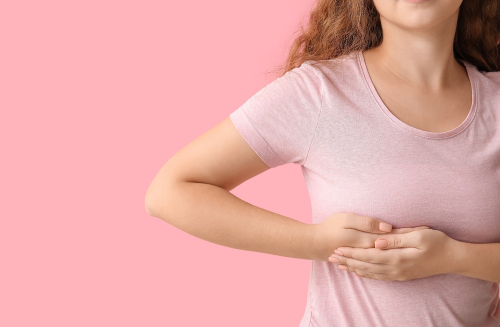 Image of a woman with her head cut off in a pink shirt with a pink background giving herself a self-breast exam.