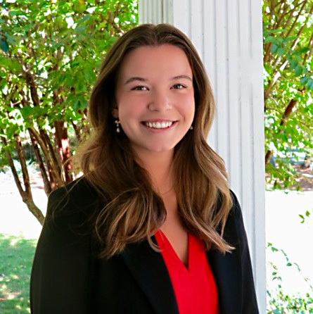 Three-quarters portrait of a white women in her 20s with brown eyes and light brown hair. She is wearing a red blouse and a black jacket. She is smiling and looking at the camera.