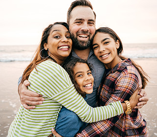 A family of four is standing closely together on a beach. They are hugging and smiling.