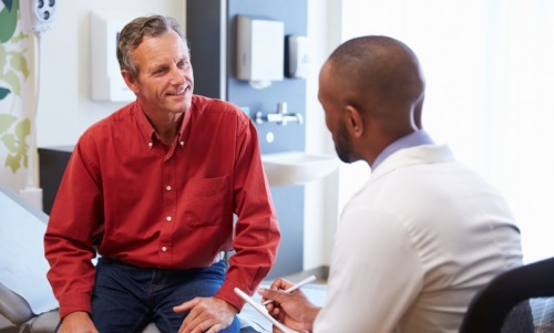 A man in a red shirt speaks to a doctor in a doctor's office.
