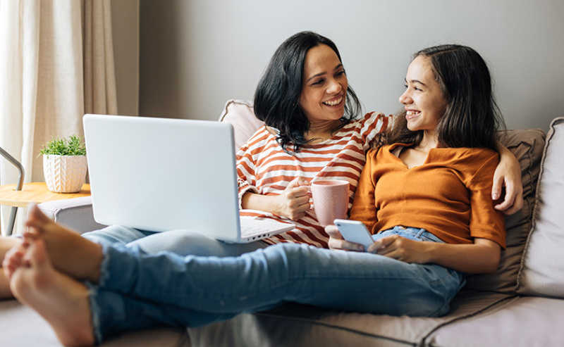 A Hispanic mother and tween daughter sit together casually on a couch. They are having a conversation with slight grins on their faces.