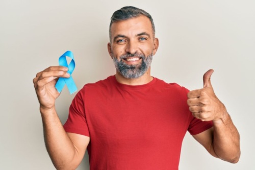 A man in a red t shirt holds a blue ribbon in his right hand and makes a thumbs up with his left while smiling.