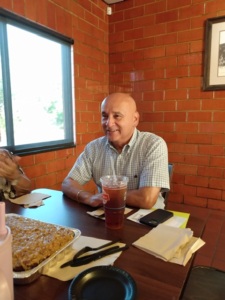 A man sits at a table with a large fountain drink, with a window and brick walls behind him.
