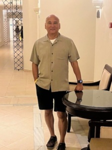 A man stands by a table wearing a tan shirt and black shorts.