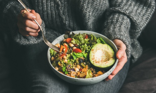 A person in a sweater holds a bowl of healthy food, including greens, grains and an avocado.