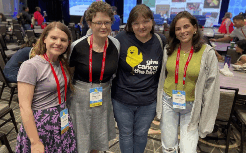 Four people pose with arms around each others shoulders, wearing red lanyards over their clothes.